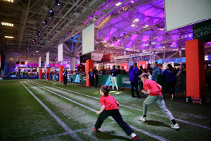 Zwei junge Jungs spielen Fußball auf einem Feld, umgeben von einer Gruppe von Menschen, mit Flaschen und anderen Gegenständen auf der rechten Seite, einem Zaun, Bannern, einem Anzeigebildschirm, Metallrahmen, einem Dach mit Deckenleuchten und Metallstangen im Hintergrund.