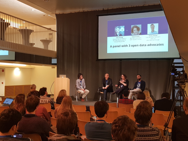 Eine Gruppe von Menschen sitzt auf Stühlen vor einem Projektionsschirm, mit einem Laptop auf einem zentralen Tisch, einer Kamera auf einem Stativ rechts daneben und Geländern, Lampen und einer Wand im Hintergrund, die an einer Podiumsdiskussion über Open-Data-Aktivisten teilnimmt.