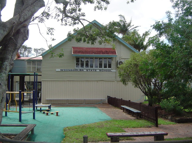 Ein Schulgebäude mit einem Spielplatz, der von einem Zaun umgeben ist, Bänken, Gras, Pflanzen, Bäumen und anderen Gegenständen, unter einem bewölkten Himmel.