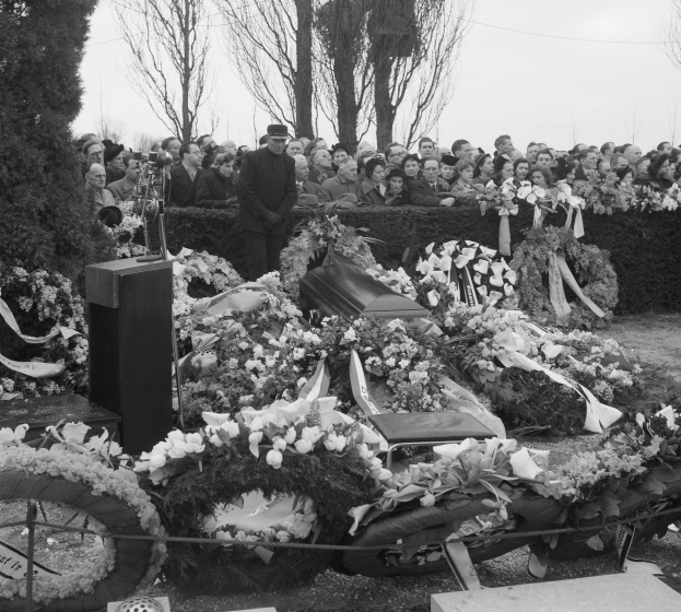 Schwarzer und weißer Trauerzug mit einem Mann in einer Mütze, der auf einem Podium spricht, umgeben von Blumen, Stühlen und einer Menge, draußen mit Bäumen und einem klaren Himmel.