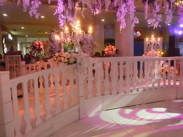 Weißer Handlauf mit Blumen und Kronleuchtern geschmückt, umgeben von Gästen, Säulen und beleuchteten Lichtern im Hintergrund sowie ein Bildschirm auf der rechten Seite des Raums.