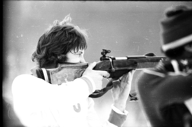 Schwarzes und weißes Bild eines Mannes in einem weißen Hemd, der mit einem Gewehr auf ein Ziel schießt und von einer anderen Person zur Rechten begleitet wird.