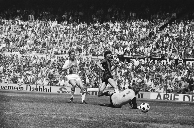 Schwarzes Foto von Männern, die auf einem Feld Füüball spielen, mit Zuschauern im Hintergrund und einer "Bundesliga 1978-79 Ajax 1-1 Hannover 96"-Plakate.