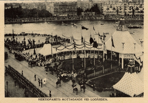 Ein altes Schwarz-Weiß-Foto zeigt eine Menge, die sich um einen Karneval mit Zelten, Fahnen, Stangen und Pferden im Vordergrund und Gebäuden, Bäumen und Wasser im Hintergrund versammelt hat, mit Text am unteren Rand des Bildes.