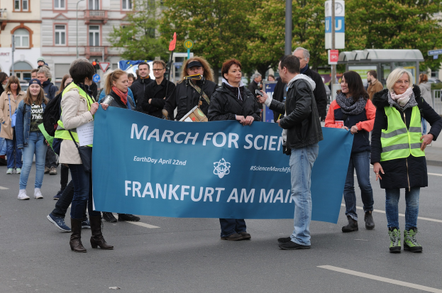 Eine bunte Gruppe von Menschen marschiert auf einer Straße, die eine "March for Science Frankfurt am Main"-Fahne trägt, mit Bäumen, Gebäuden und einem klaren Himmel im Hintergrund.