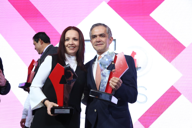 Zwei strahlende Menschen auf einer Bühne mit Trophäen und einem Bildschirm im Hintergrund