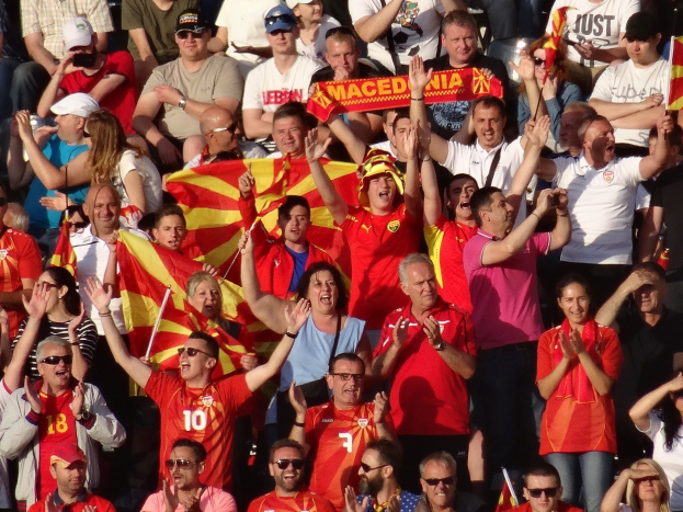 Eine Gruppe von Menschen in einem Stadion, die ihre Hände heben, einige tragen Kappen und Schutzbrillen und halten Fahnen und Schilder mit Text, mit Geländern im Hintergrund.