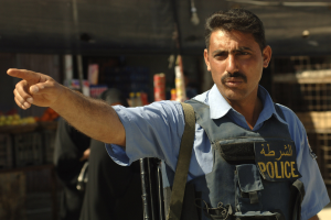 Polizeibeamter in Uniform mit Pistole vor einem Geschäft stehend, mit unscharfen Personen und Gegenständen im Hintergrund.