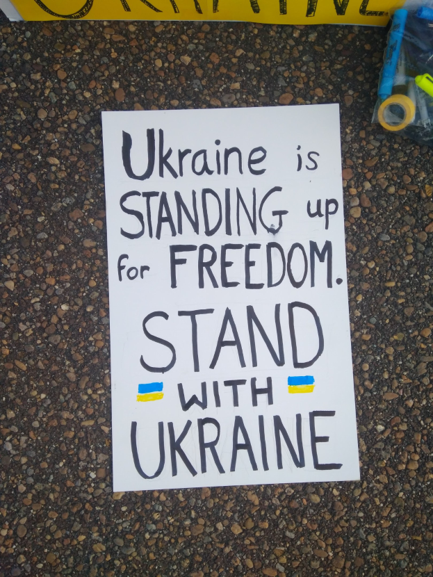 Ein weißes Papier mit der Aufschrift "Ukraine steht für Freiheit auf, steh auf für Ukraine" liegt vor einer gelben Fahne auf dem Boden. Rechts daneben befindet sich eine Plastikabdeckung mit verschiedenen Gegenständen.