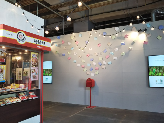 Ein Ladeneinrichtungsbild mit einer Auslage voller verschiedener Lebensmittel, einem roten Briefkasten auf dem Boden, einem Display auf der Wand mit einem Yokohama Kitchen-Schild, Papieren an den Wänden, Deckenleuchten und sichtbaren Rohren.