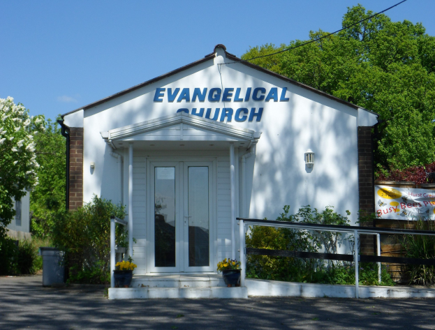 Evangelische Kirche mit Straße davor, umgeben von Grün und einem klaren blauen Himmel.