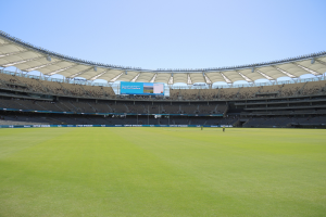 Ein großes Stadion mit einem grünen Feld, umgeben von Zuschauern, Werbetafeln und einem großen Bildschirm oben, unter einem sichtbaren Himmel.