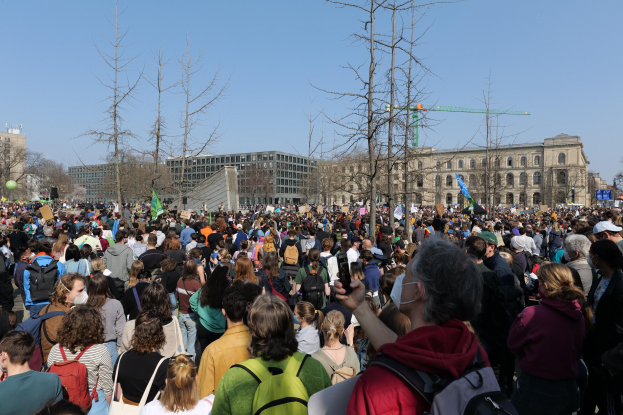 Eine große Menge Menschen mit Schildern und Taschen steht vor einem Gebäude mit Bäumen und einem klaren Himmel während einer Klimaprotest.