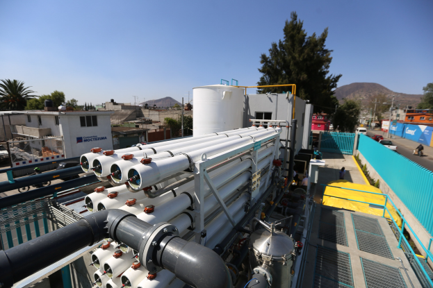 Große Gruppe von Rohren auf einem Gebäudedach mit Industriegeräten, Fahrzeugen, Menschen, Gebäuden, Bäumen und einem klaren blauen Himmel.