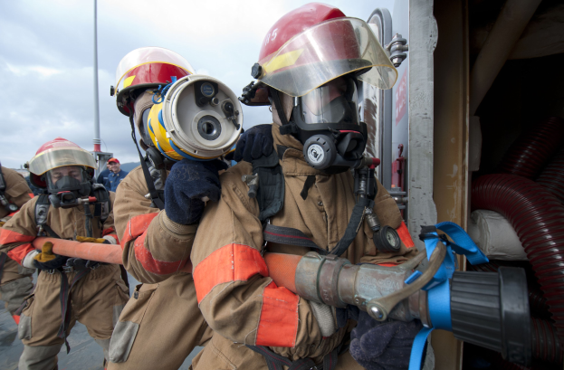 Feuerwehrleute in Schutzausrüstung mit einem Schlauch haltenden, vor einem Mast und Rohren gegen einen bewölkten Himmel stehend.