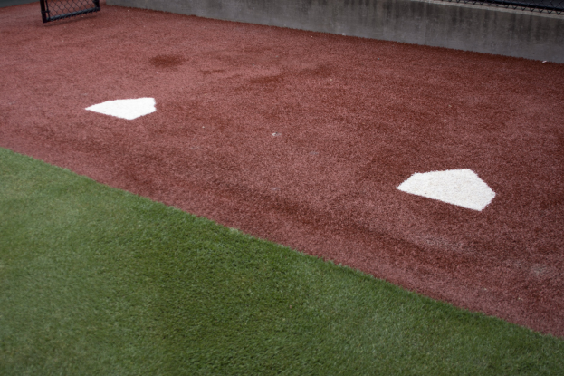 Ein Baseballfeld mit Kunststoffrasen, einem Zaun, einem Home Plate in der Mitte und einer Wand im Hintergrund.