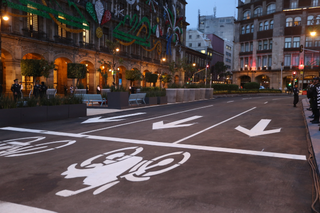 Eine Stadtstraße mit einem Fahrradweg, Gebäuden, Straßenmasten, Straßenlaternen, Ampeln, Menschen auf dem Gehweg, Bänken, Pflanzen, Bäumen, dekorativen Gegenständen und einem klaren blauen Himmel.