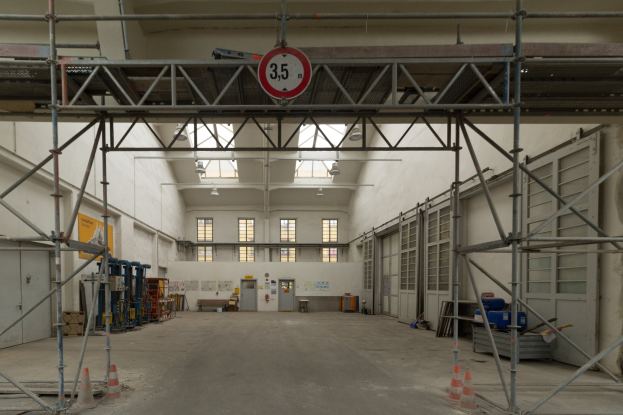 Großes Lagerinnere mit ausgedehnter Gerüstkonstruktion, Verkehrskegeln, Metallstangen, einem Schild, Maschinen, einer Tür, Fenstern, Wänden, Deckenleuchten und Dach.