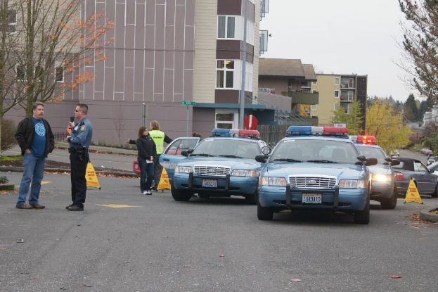Autos auf einer Straße mit vier Menschen in der Nähe, Gebäuden mit Fenstern im Hintergrund, Bäumen und einem Notfallwarnschild.
