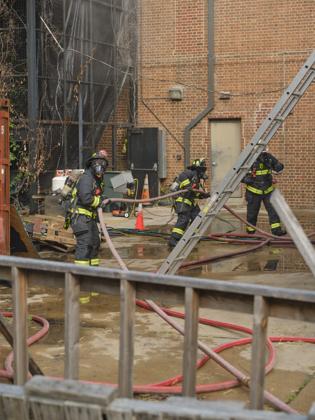 Feuerwehrleute mit Helmen arbeiten daran, ein Gebäude Feuer zu löschen, umgeben von Ausrütung und einem Metallzaun, mit einem Baum und Himmel im Hintergrund.