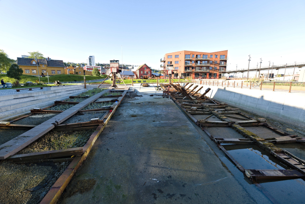 Baustelle mit Stehender Wasser auf einer Straße mit Holzbohlen, neben einer Wand, Bäumen und Gebäuden unter einem Himmel.