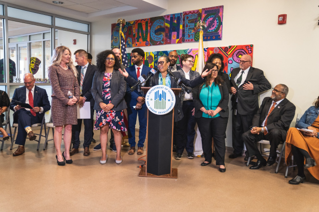 Eine Gruppe von Menschen, die vor einem Podium mit Mikrofon und einer Tafel mit der Aufschrift "Sancho-Stadtrat tritt dem Stadtrat bei" stehen und sitzen, mit Flaggen, Feuerlöscher und Deckenleuchten an der Rückwand.