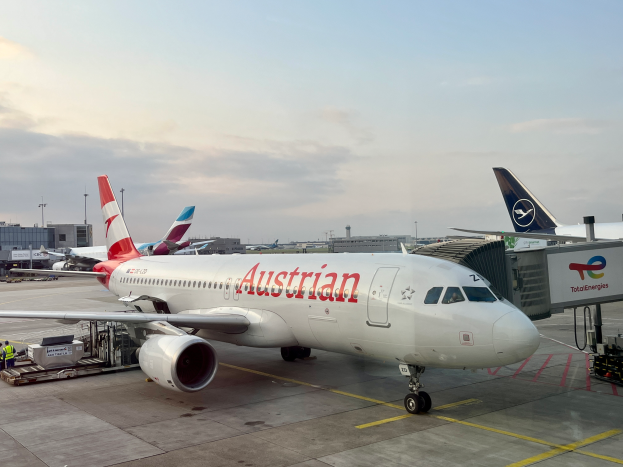 Ein Airbus A330-300 der Austrian Airlines auf dem Rollfeld am Frankfurter Flughafen, umgeben von Menschen, Fahrzeugen und anderen Gegenständen, mit Gebäuden, Masten und einem klaren blauen Himmel im Hintergrund.