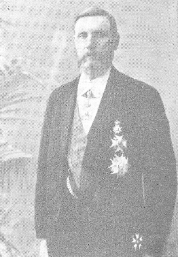 Ein Mann in einem schwarzen und weißen Anzug und Krawatte steht mit einem ernsten Gesichtsausdruck vor einer üppigen grünen Pflanze.
