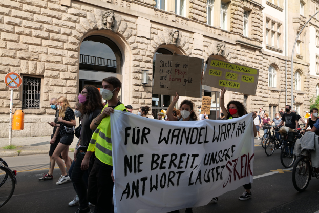 Eine Gruppe von Menschen, die während einer Demonstration die Straße entlanggehen, einige halten Schilder und andere fahren Fahrräder, mit einem Gebäude im Hintergrund, das Fenster, Bögen, Säulen und Skulpturen aufweist.