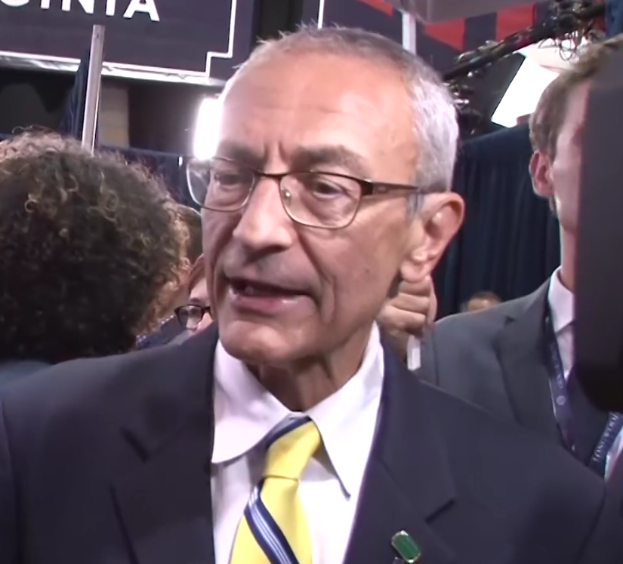 Ein Mann im Anzug und Krawatte, der eine Brille trägt, spricht zu Reportern mit einem Mikrofonständer vor ihm, umgeben von einer Gruppe von Menschen, mit Lampen und einer Tafel im Hintergrund.