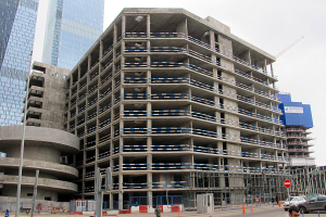 Großer Bau in einer Stadt mit Wolkenkratzern, Fahrzeugen, Fußgängern, Verkehrs-Infrastruktur und Kränen vor einem klaren Himmel.