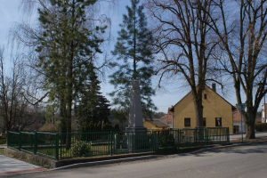 Ein Holocaust-Denkmal steht in der Mitte einer Straße, umgeben von einem Metallzaun, umgeben von Grün, Häusern und einem bewölkten Himmel.