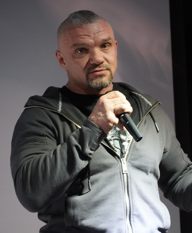 Ein Mann in einer schwarzen Kapuzenjacke und schwarzem Anzug mit einem weißen Hemd, der ein Mikrofon hält, steht selbstbewusst vor einer Wand und scheint ein Komiker zu sein.