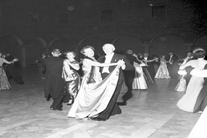 Schwarzes und weißes Foto einer Gruppe von Menschen in Abendgarderobe, die in einem Ballsaal tanzen, mit einer Wand im Hintergrund.