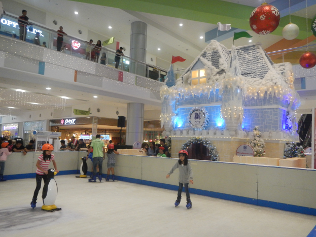 Eine Gruppe von Menschen, die auf einer Eisbahn in einem Einkaufszentrum Schlittschuh laufen, umgeben von einer hauseigenen Struktur, die mit Lichtern und festlichen Dekorationen geschmückt ist, mit Zuschauern und zusätzlichen Dekorationen, die eine feierliche Atmosphäre schaffen.