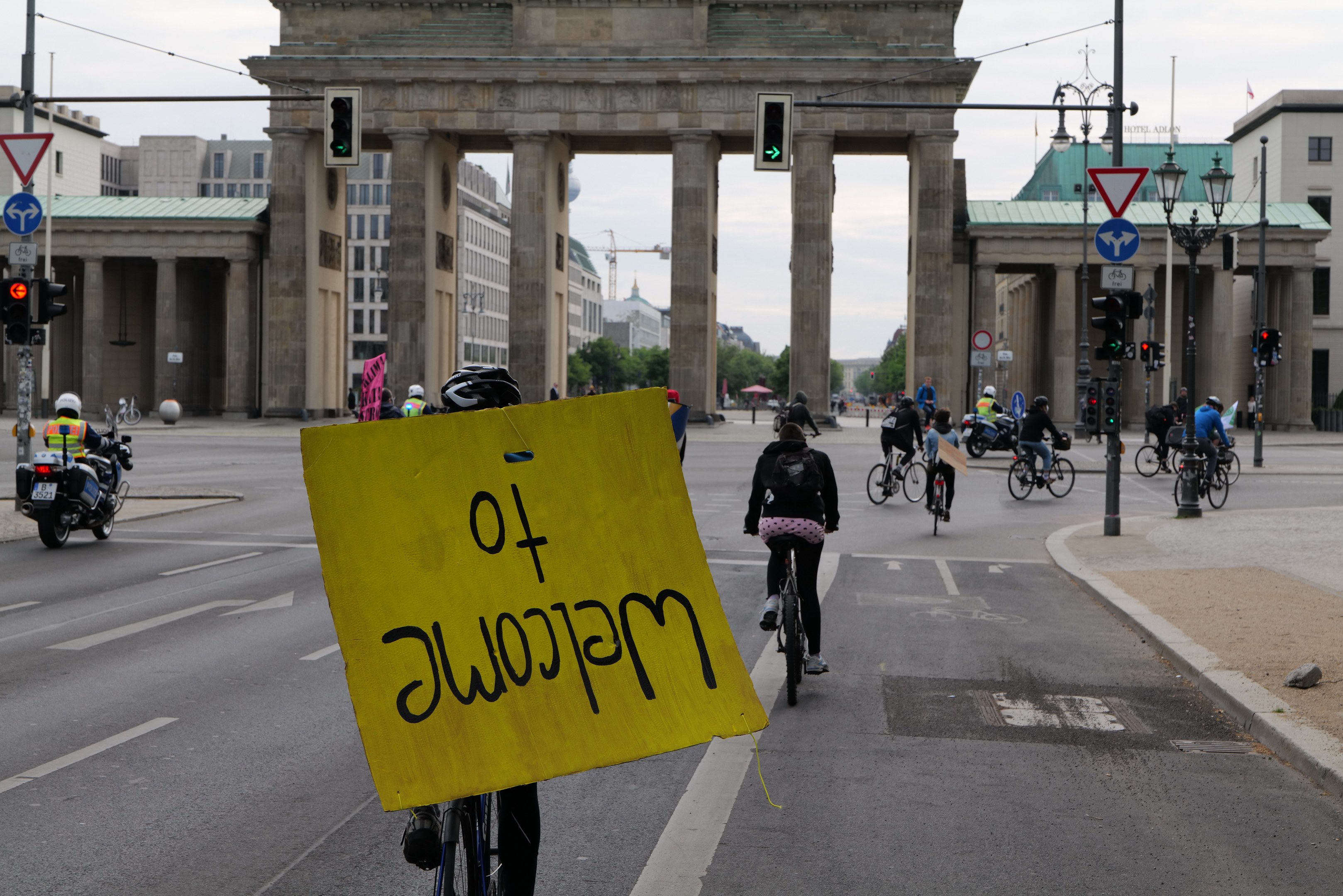 Eine Gruppe von Radfahrern fährt am Brandenburger Tor in Berlin vorbei, einer hält ein gelbes Schild, mit Laternenpfählen, Ampeln, Gebäuden, Bäumen und einem klaren Himmel im Hintergrund.