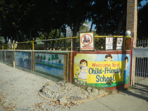Ein Wandschild mit der Aufschrift "Willkommen in einer kinderfreundlichen Schule" vor einem Metallzaun, Bäumen, Steinen und einem Weg unter einem bewölkten Himmel.