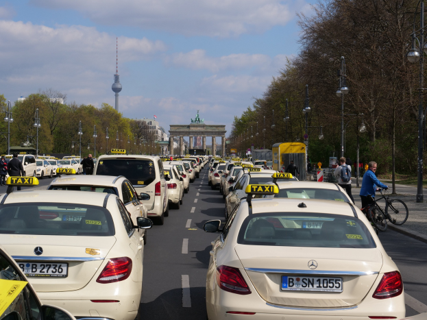 Eine lange Reihe von Taxis, die entlang einer belebten Straße in Berlin, Deutschland, geparkt sind, mit Fahrrädern und Fußgängern auf dem Gehweg, flankiert von Laternenmasten und Bäumen und Gebäuden, einem Bogen und einem Turm im Hintergrund unter einem bewölkten Himmel.
