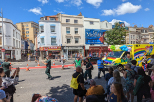 Eine Gruppe von Menschen vor einem Rettungswagen auf einer Straße mit Absperrgittern, Bäumen, Gebäuden mit Fenstern, Laternen und einem Banner mit Text, unter einem bewölkten Himmel bei der London Pride Parade.