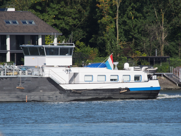 Ein großes Boot fährt einen Fluss hinunter, mit einem Haus auf der linken Seite und Bäumen im Hintergrund.