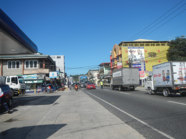 Eine belebte Stadtstraße mit Fahrzeugen, Fußgängern, Gebäuden, Schildern, Strommasten, Bäumen und einem bewölkten Himmel.