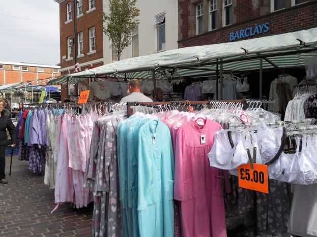 Ein Flohmarkt mit Kleidung auf Ständern, einschließlich Kleider an Bügeln mit Preisschildern, Menschen, die Stände durchsuchen, und Gebäude, ein Baum und ein klarer blauer Himmel im Hintergrund.