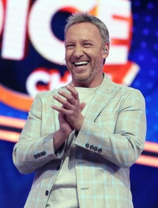 Ein Mann in einem Anzug, der auf einer Bühne klatscht, mit dem 'The Voice'-Logo im Hintergrund.