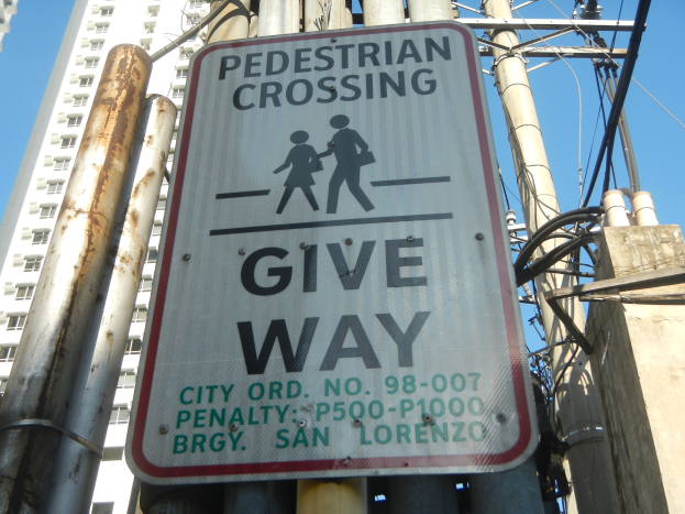 Ein am Mast angebrachtes Schild mit der Aufschrift "Fußgängerübergang Vorrang" vor elektrischen Masten und Drähten, einem Gebäude und dem Himmel.