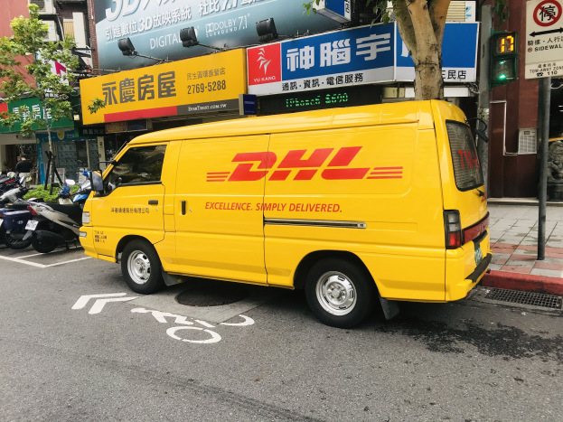 Gelber DHL-Lieferwagen parkt am Straßenrand, umgeben von Gebäuden, Bäumen, Schildern, Strommasten, Verkehrszeichen und einem Fußgänger auf dem Gehweg.