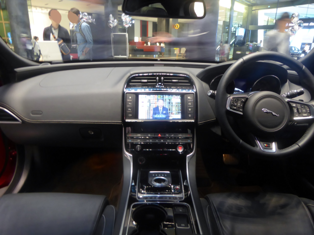 Innenansicht eines Jaguar F-Pace R-Dynamic S AWD mit Lenkrad, Armaturenbrett, Bildschirm, Knöpfen, Sitzen, Spiegel und Glasfenstern. Im Hintergrund sind Menschen, Lichter und andere Gegenstände durch die Fenster zu sehen.