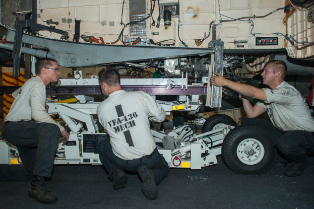 Drei Männer in Uniformen arbeiten an einem Flugzeug in einer Halle, umgeben von Werkzeugen und Equipment, scheinbar um es zu reparieren.