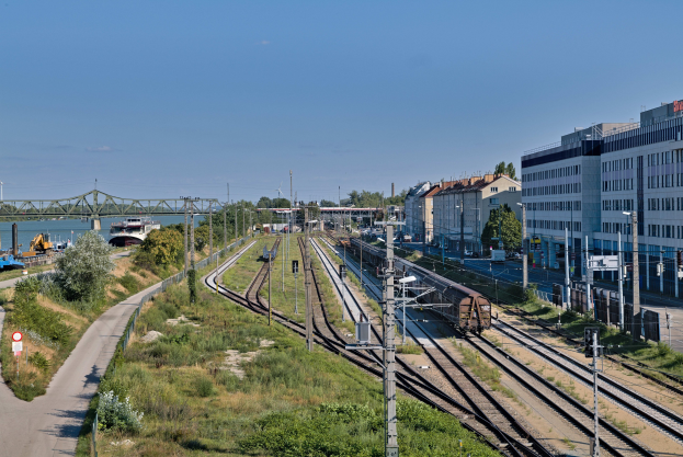 Ein Zug fährt auf Schienen neben einer Stadtlandschaft mit Infrastruktur und Grün unter einem bewölkten Himmel.