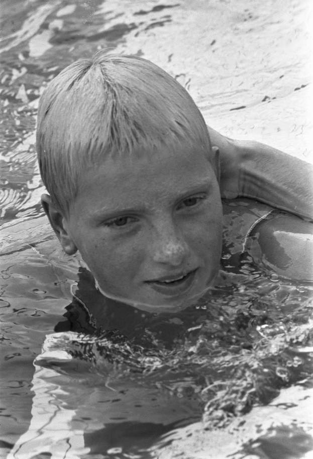Ein junger Junge schwimmt in einem Pool mit ausgestreckten Armen, sein Gesicht von der Sonne beleuchtet, während er durch das Wasser gleitet, die Augen geschlossen und das Haar nass.