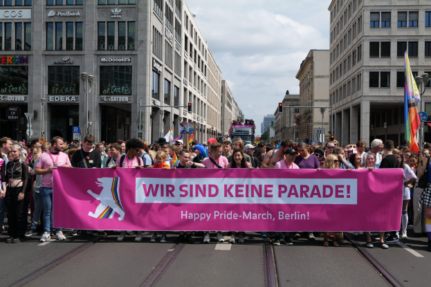 Eine Gruppe von Menschen marschiert auf einer Berliner Straße und hält ein pinkes Banner mit der Aufschrift "Happy Pride March", während Gebäude, Laternenpfähle und Verkehrszeichen die Straße säumen und der Himmel bewölkt ist.